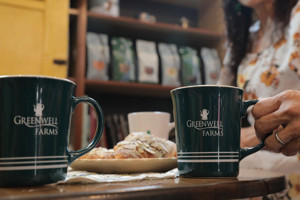 Two Greenwell Farms branded coffee mugs on a wooden table inside a cozy coffee shop setting