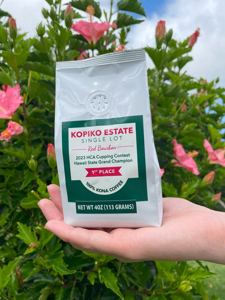 A hand holding a bag of Kopiko Estate Red Bourbon 100% Kona Coffee in front of pink flowers